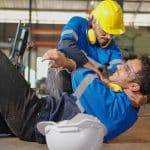 Injured construction worker laying on the ground in pain being held up by his coworker